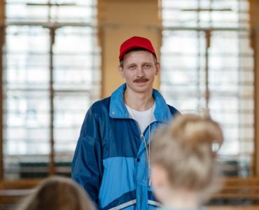 Male coach in a gym engaging with a group of children, wearing casual sportswear.